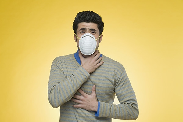 Header image showing a person with a pollution mask covering his mouth and nose. He's grasping at his throat with his right hand. He's wearing a sweater with horizontal stripes (blue and yellow). The image has a yellow background.