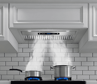 A frontal-bottom shot of the IKTCH IKB02-30 (30) insert range hood. Its brushed stainless steel in color. It's integrated into a white cabinet. There are two pots under it on a stove with turned on burners. There is steam coming off of the pots which is sucked up by the range hood.