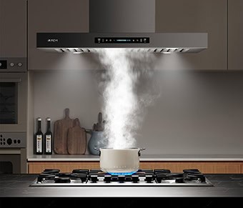A kitchen shot of the IKTCH IKIS02-36-M island range hood. It's brushed stainless steel in color and has a touch screen and baffle filters. It has a pot under it with steam coming off of it. It's a dark kitchen and the stove it lit by the LED lighting.