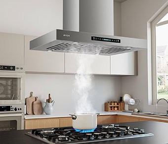 A kitchen shot of the IKTCH IKIS02-36-M island range hood. It's brushed stainless steel in color and has a touch screen and baffle filters. It has a pot under it with steam coming off of it. The cupboards behind it are white.